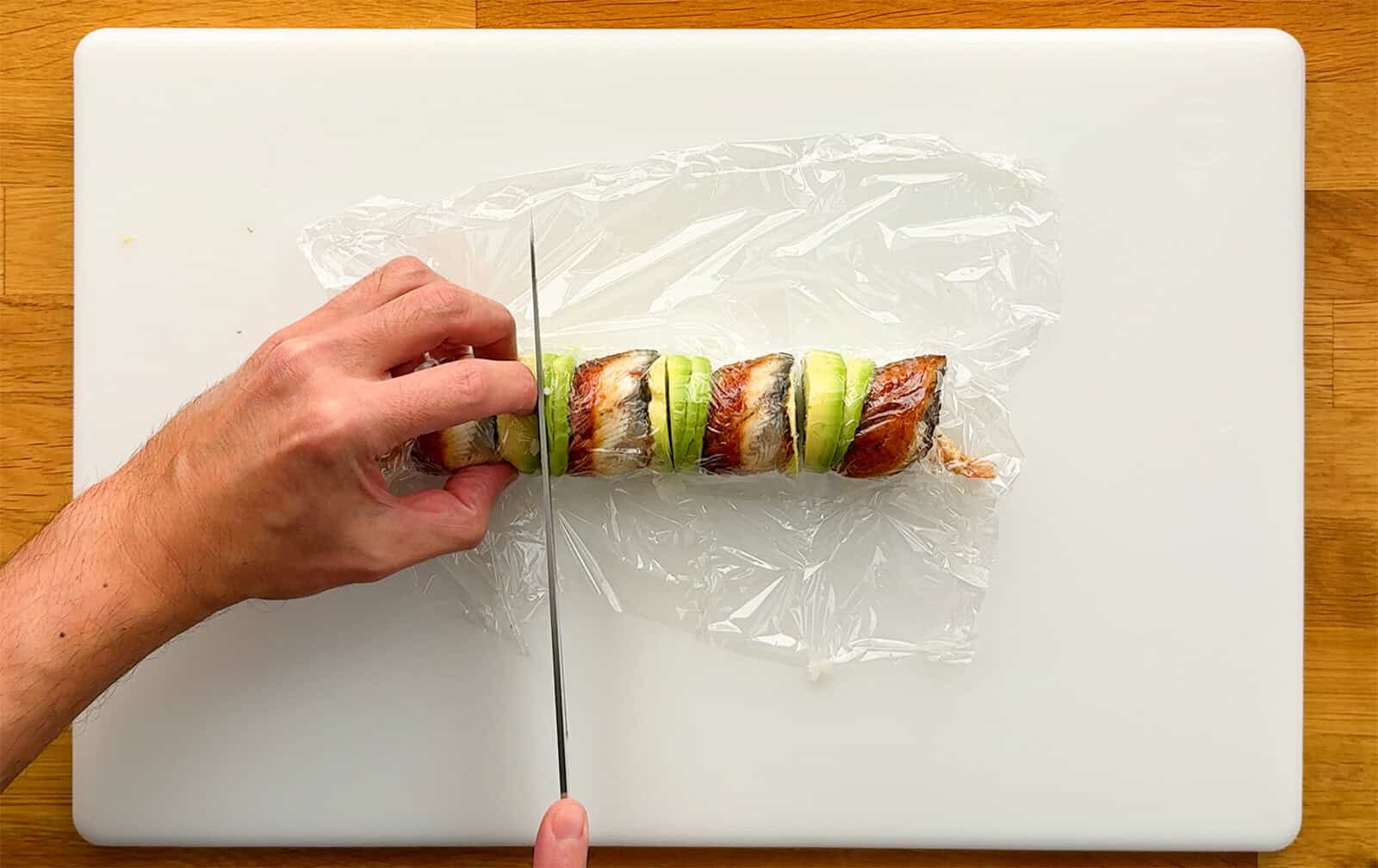 Cutting a Dragon Roll into pieces with a knife while keeping the plastic wrap in place.