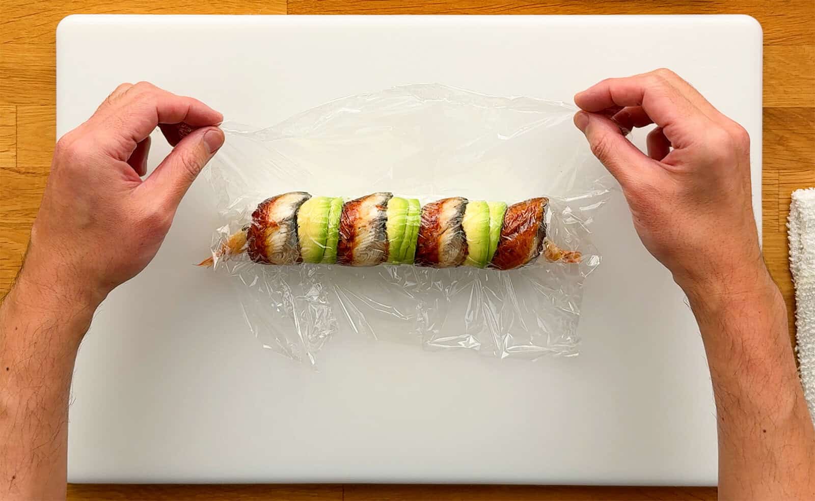 Hands holding the excess plastic wrap above a Dragon Roll after shaping the unagi and avocado toppings.