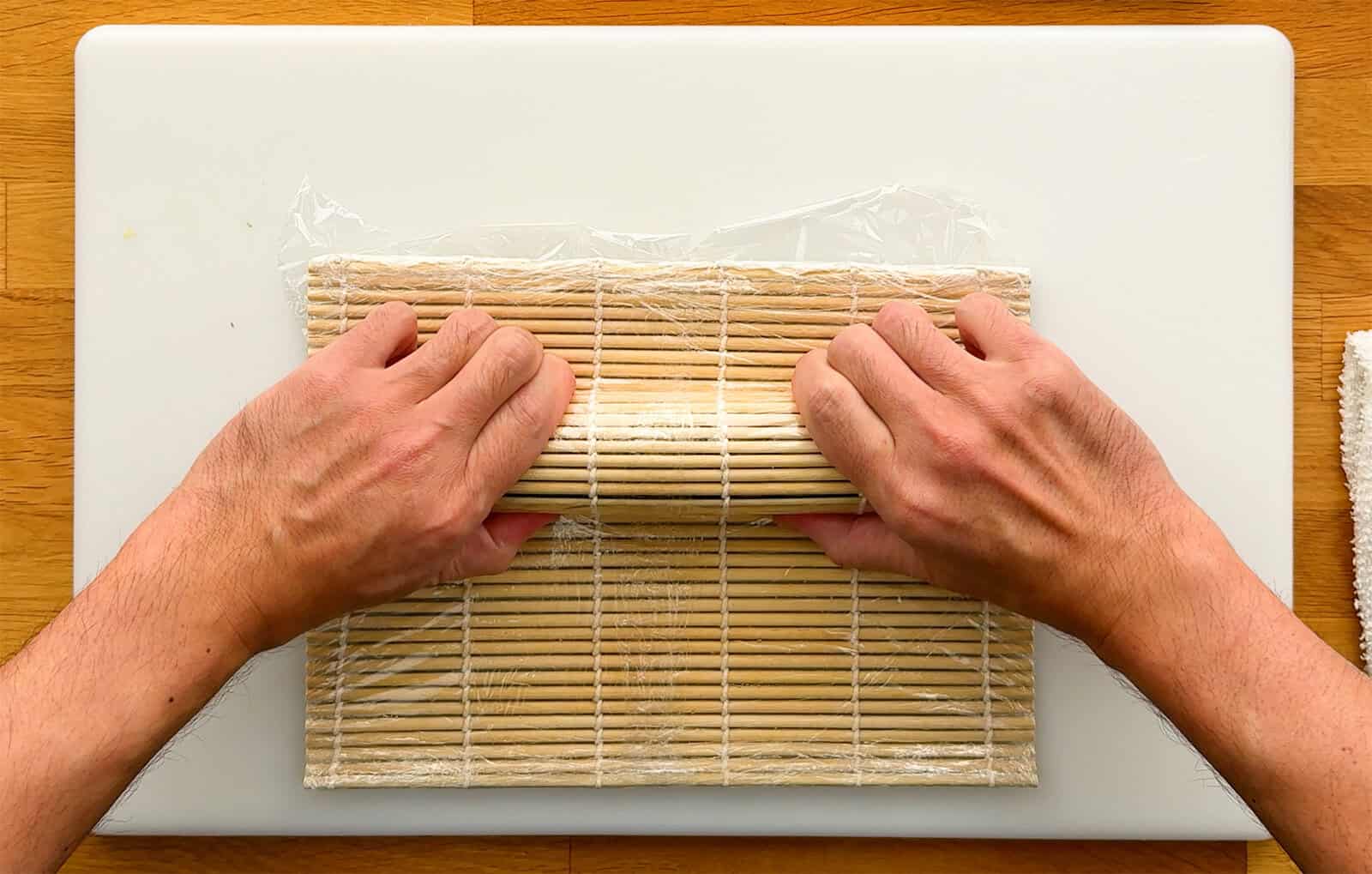Hands using a bamboo sushi rolling mat to shape the unagi and avocado toppings on a Dragon Roll.