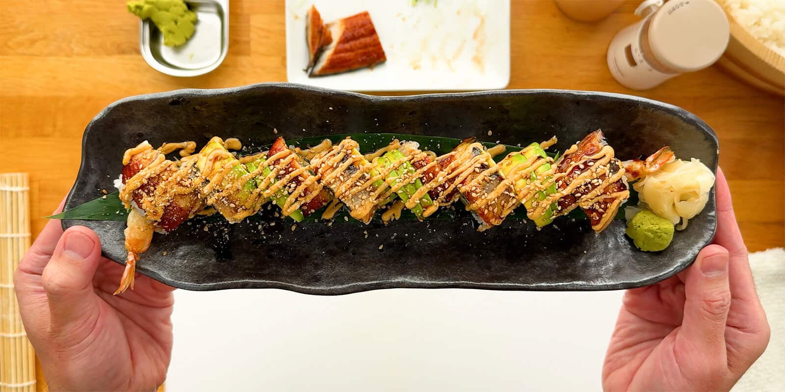 Homemade Dragon Roll sushi topped with unagi, avocado, spicy mayo, unagi sauce, and sesame seeds, served on a black plate.