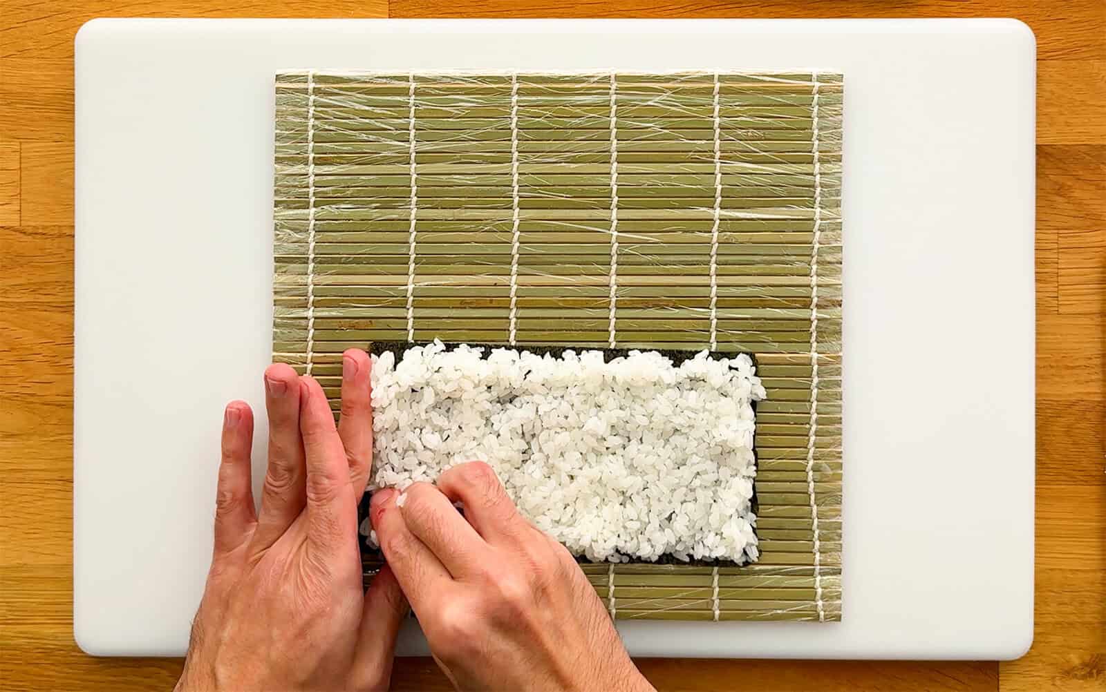 Spreading sushi rice evenly over a half sheet of nori on a bamboo rolling mat